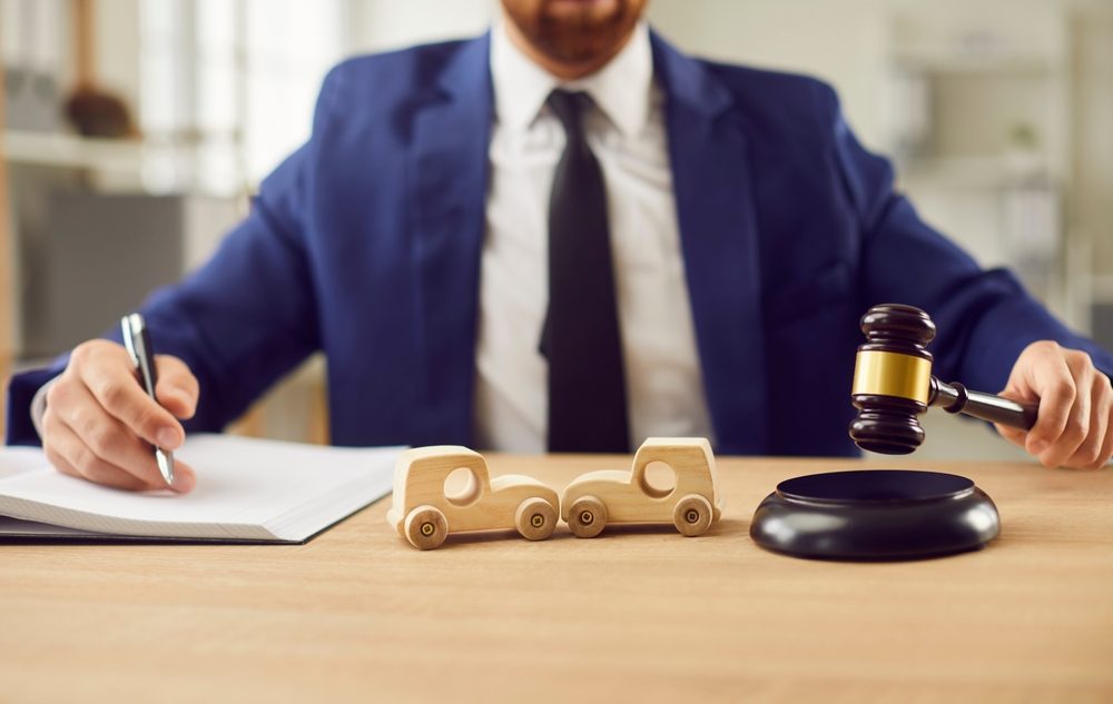 Two wooden car toys on a table in front of a lawyer holding a judge’s gavel who is meeting with someone looking to find a lawyer for a car accident.